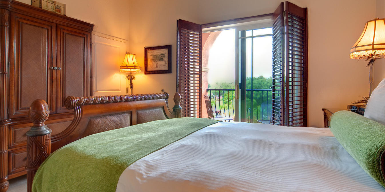 A bright hotel-style bedroom with a wooden bed, green blanket, shutters, balcony door, and bedside lamps.