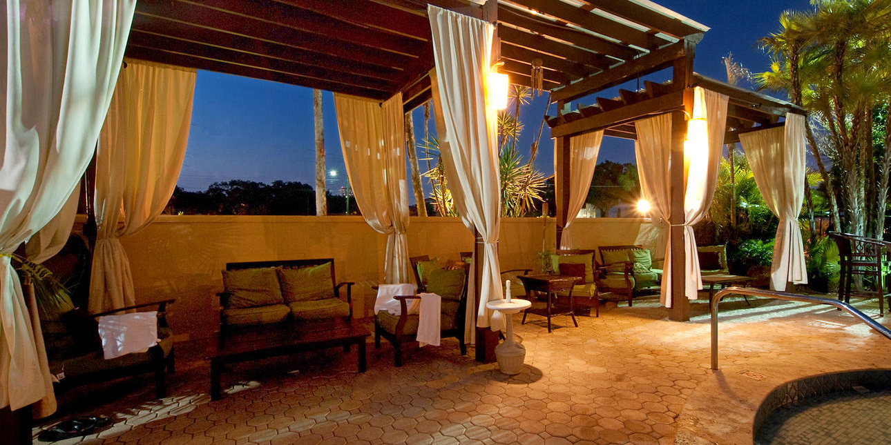 An outdoor patio at dusk with a wooden pergola, billowing white curtains, warm lights, lush palms, and a small pool.