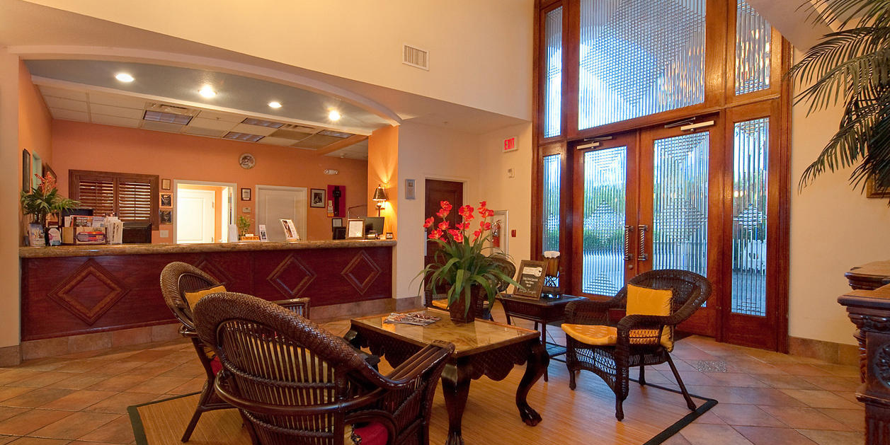 A warm hotel lobby with a wooden reception desk, wicker chairs, tiled floors, and large arched windows with orchids.