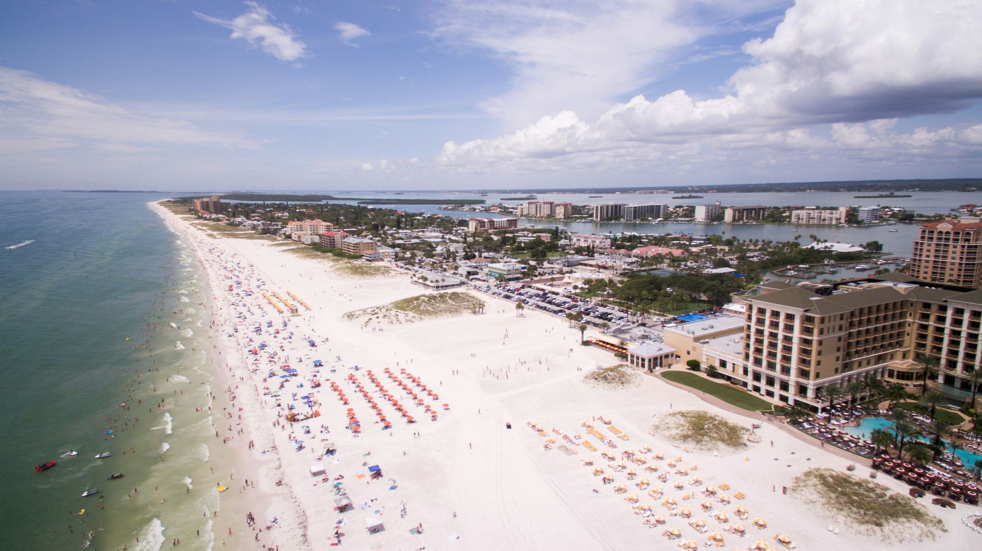 Aerial view of a crowded sandy beach with umbrellas, boats, and beachfront hotels along a calm turquoise coastline.