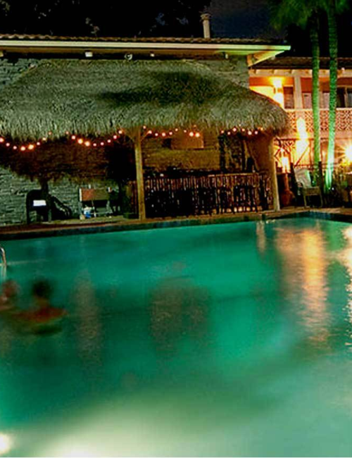Nighttime resort pool with lit string lights, loungers, thatched cabanas, and a building in the background.
