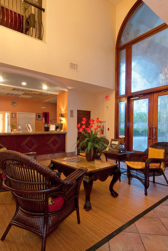 A hotel lobby with a reception desk, wicker chairs, a wooden table, and a vase of red orchids by large arched windows.