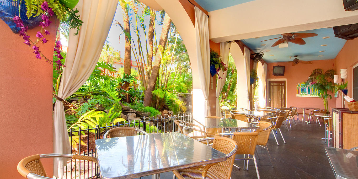 A tropical outdoor patio with wicker chairs and tables, tall palm plants, hanging flowers, and ceiling fans under a bright sky canopy.