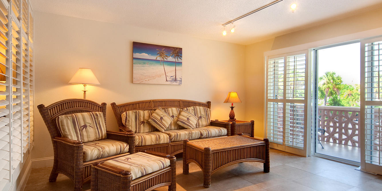 A sunny beach-themed patio living room with wicker seating, lamps, wall art, and glass doors opening to a sunny balcony.