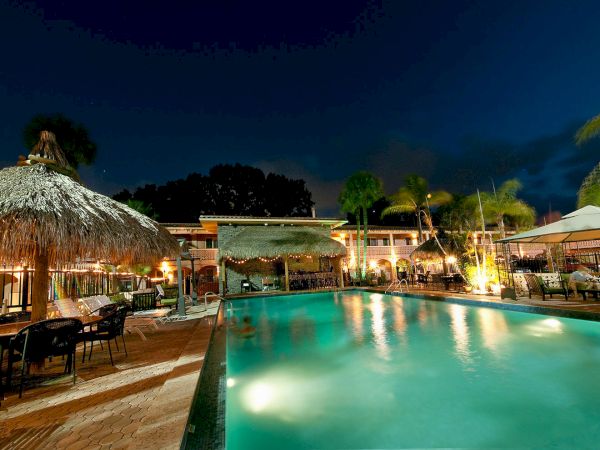 A lit outdoor resort pool at night with palm trees, tiki huts, and lounge chairs under a dark blue sky.