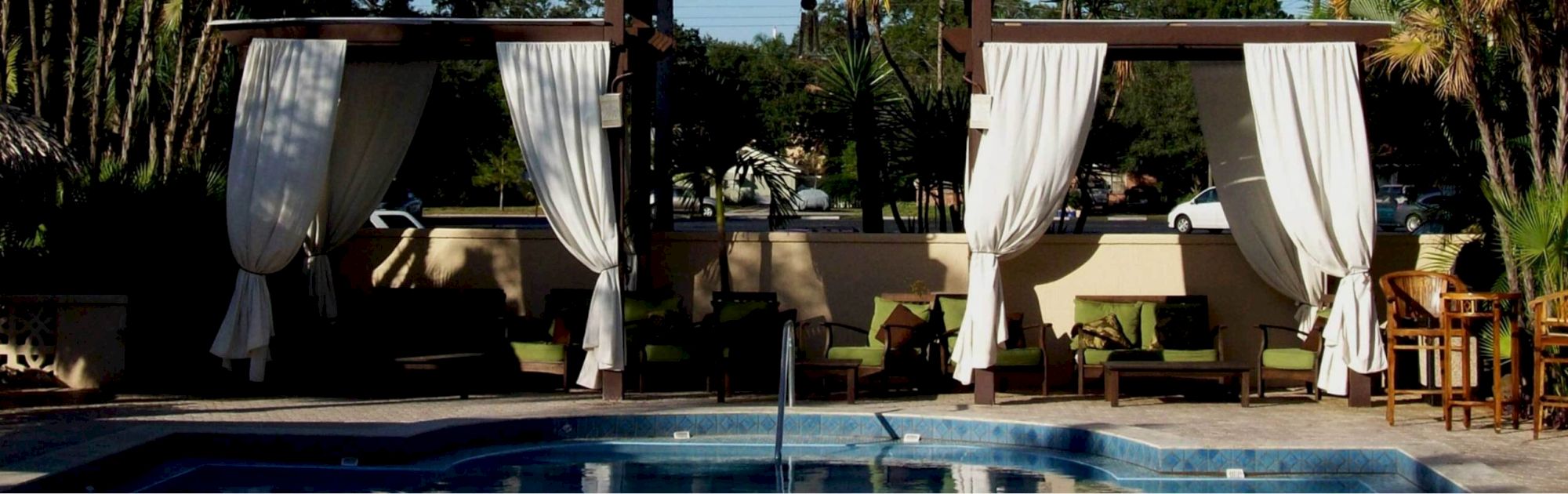An outdoor pool with a shaded cabana, billowing curtains, lounge chairs, and palm trees in the background.