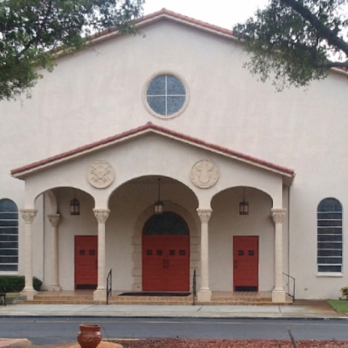 White stucco church with three red doors and arches, blue shutters, and trees in front.