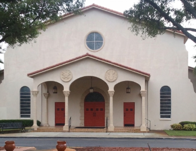 White stucco church with three red doors and arches, blue shutters, and trees in front.