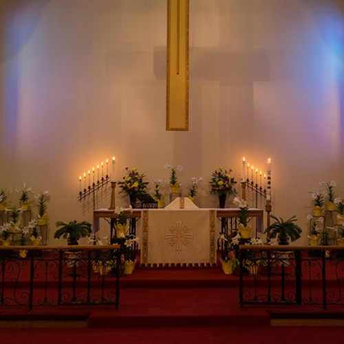 A church altar decorated with flowers and lit candles, framed by stained-glass blue window light.