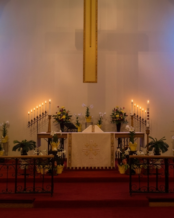 A church altar decorated with flowers and lit candles, framed by stained-glass blue window light.