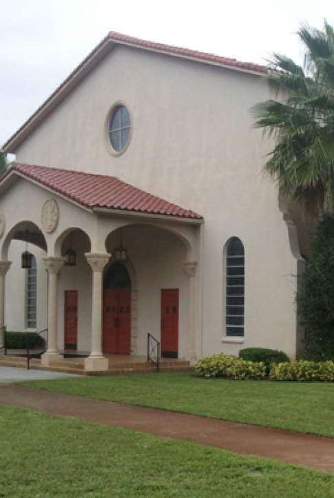 A white church building with arched windows and red doors, surrounded by trimmed shrubs and a &ldquo;Good Shepherd&rdquo; sign.