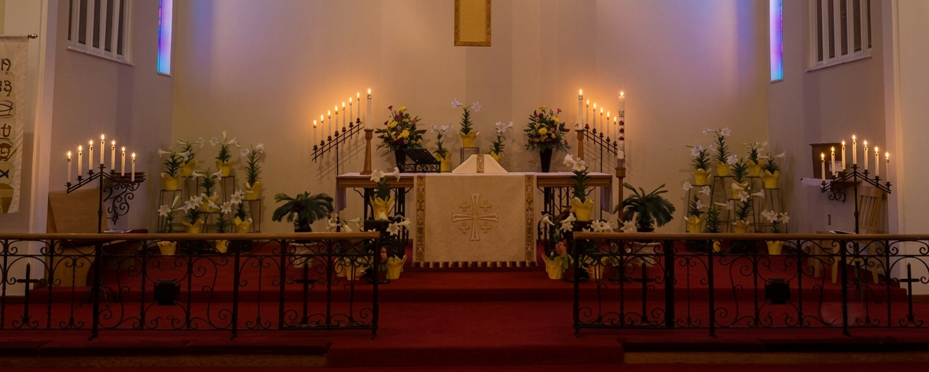 A decorated church altar with many lit candles and flower arrangements beneath blue-lit stained-glass windows.
