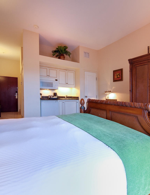 An upscale hotel bedroom with a carved wooden headboard, green-and-white bedspread, bedside lamps, and a small kitchenette in view.