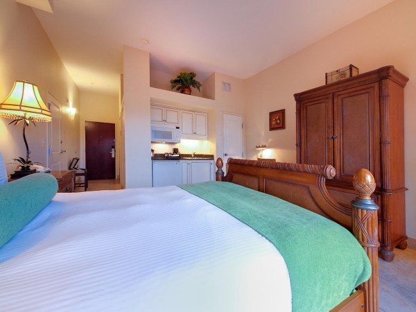 A neatly made hotel bedroom with a wooden headboard and lamps, plus a kitchenette and a large armoire in the background.