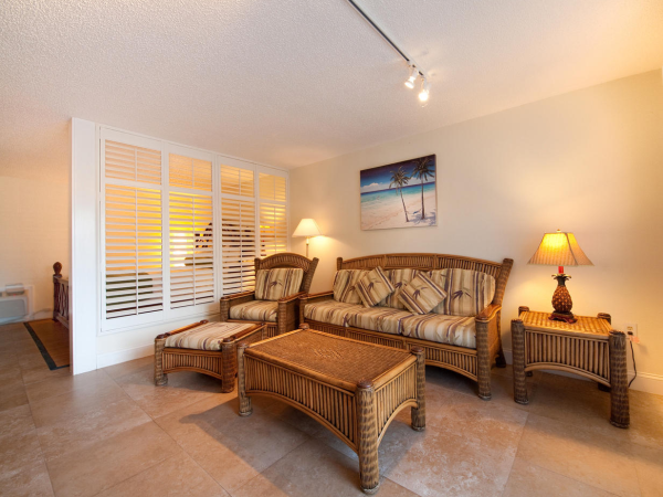 A sunny living room with wicker sofa and chairs, striped cushions, a beach wall painting, and lamps.