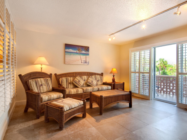 A bright beach-themed living room with wicker sofa and ottoman, lamps, wall art, and sliding doors to a balcony.
